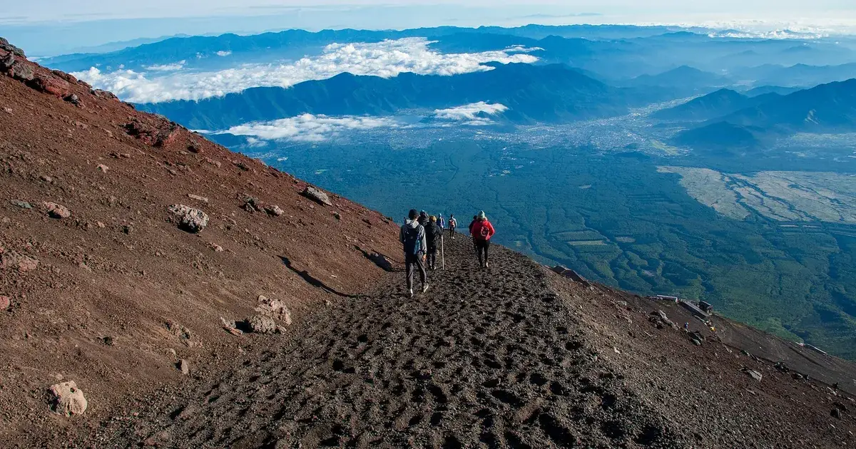 Climbers walking down the dark volcanic cinder of the Yoshida Trail on Mount Fuji in early-morning light, with the Fujiyoshida valley, forested ridges and low cloud far below