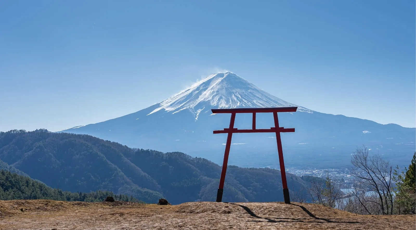 A lone red torii gate on a ridge with snow blowing from Mount Fuji's summit