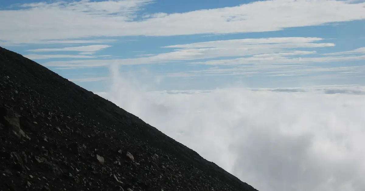 The dark volcanic scree of Mount Fuji's upper slopes dropping away above a sea of cloud under a blue summer sky