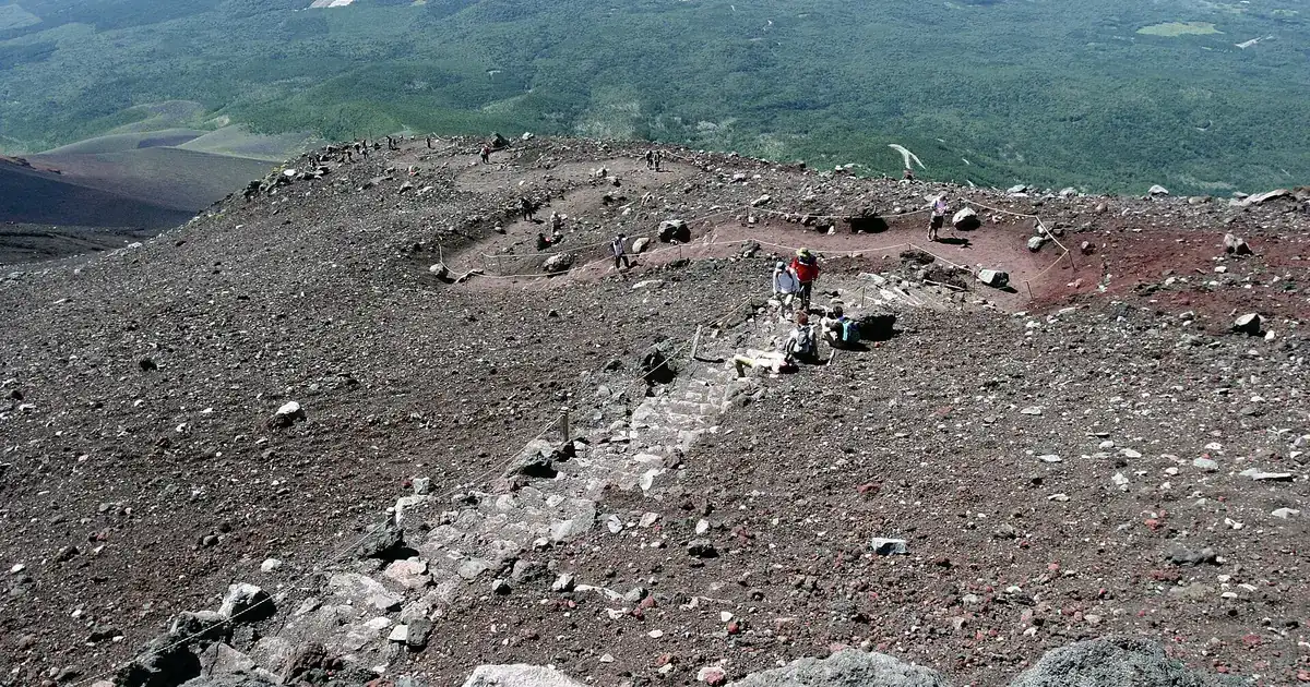 Looking down Mount Fuji's upper slopes from the 9th Station: a stepped path of red volcanic rock, climbers resting beside it, and the forested lower flanks spread out far below