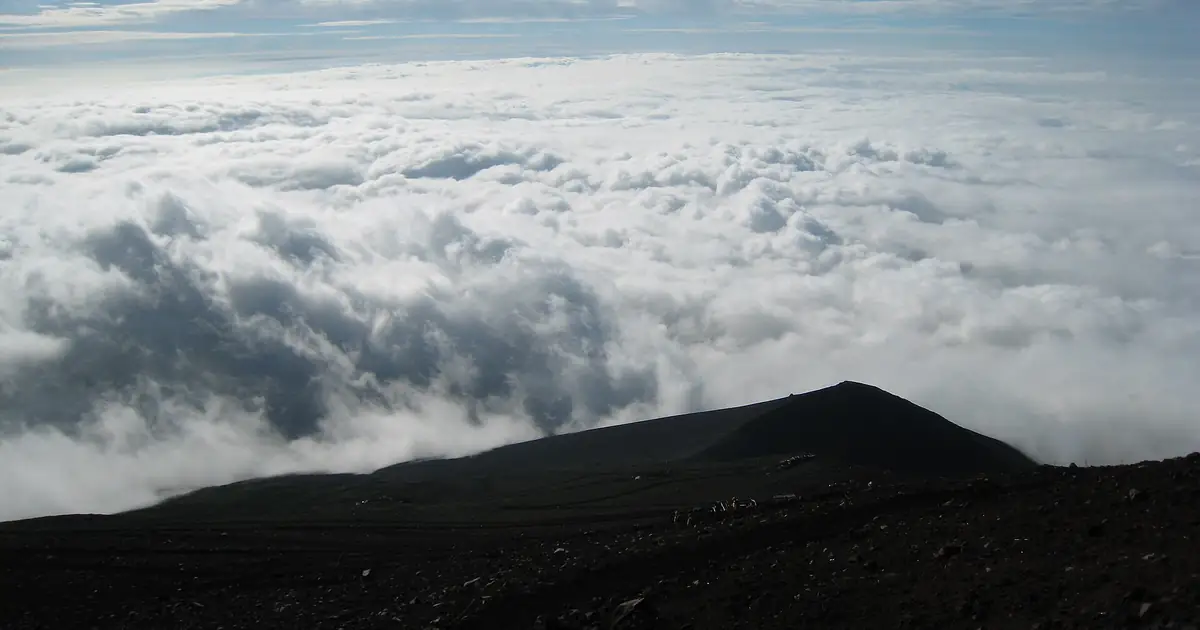 A sea of cloud seen from Mount Fuji's upper slopes, with a dark volcanic ridge in the foreground under a blue summer sky